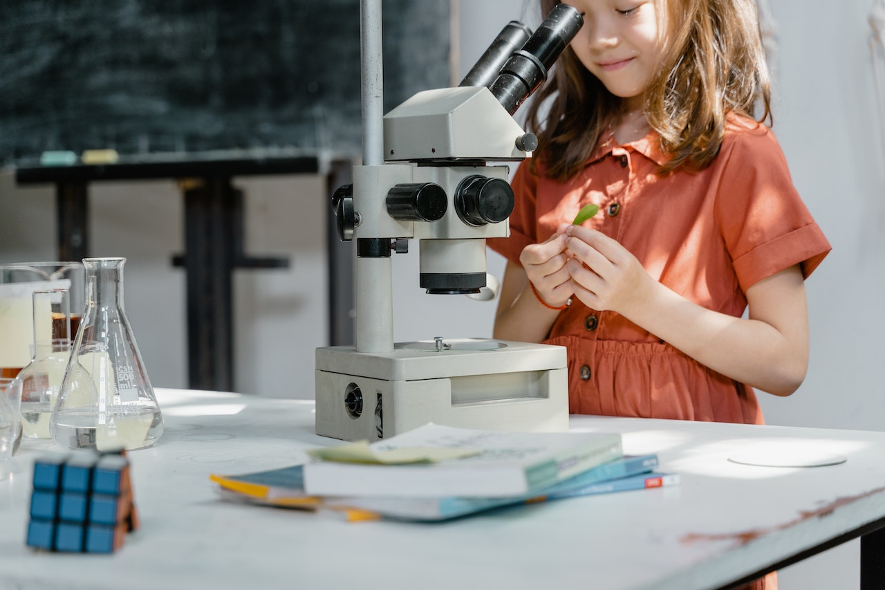 A young girl in an orange dress is standing beside a microscope on a table with books and laboratory glassware. She is holding a green leaf, appearing to focus on it. There is a Rubik's cube and a chalkboard in the background.